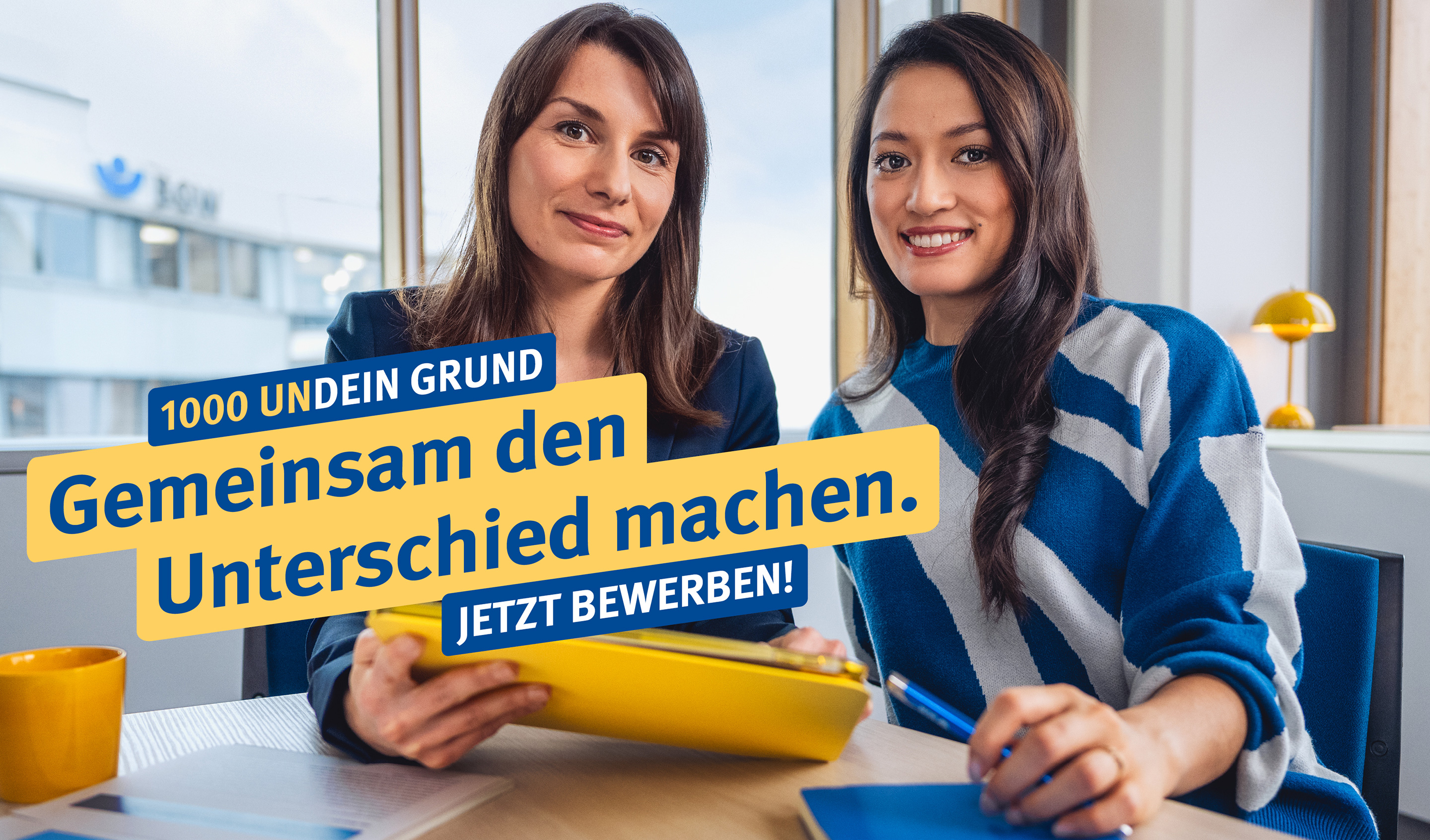 1000UNDEIN GRUND Zwei junge Frauen sitzen nebeneinander vor einem Fenster im Büro und lächeln in die Kamera.