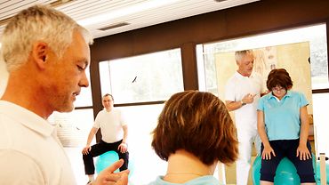 Physiotherapieraum: Physiotherapeut erklärt einer Patientin etwas vor einem Spiegel. Im Hintergrund trainiert ein weiterer Patient.