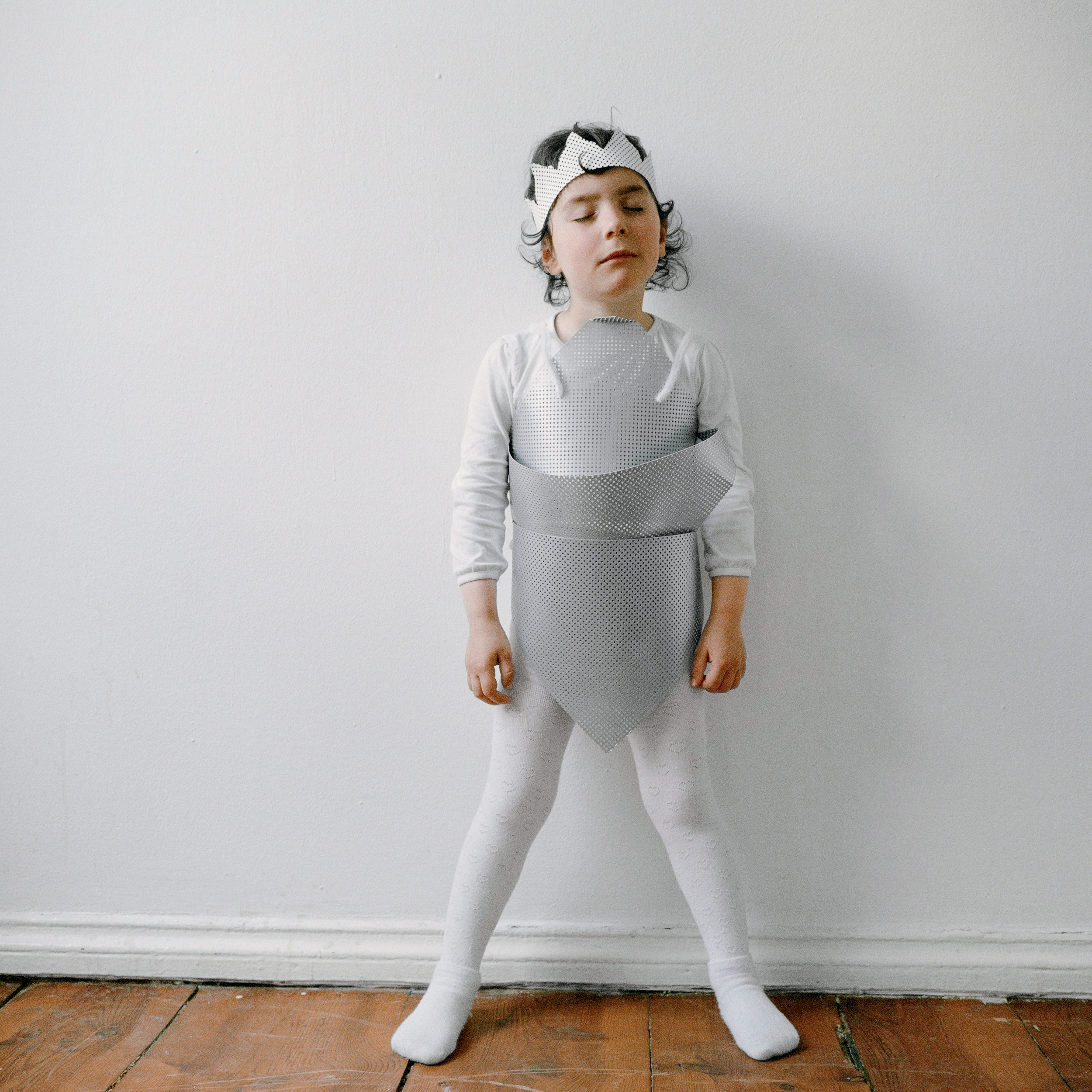 Ein kleines Mädchen steht in Strumpfhose und selbstgebastelter grauer Rüstung und mit einer grauen Krone auf dem Kopf vor einer weißen Wand.