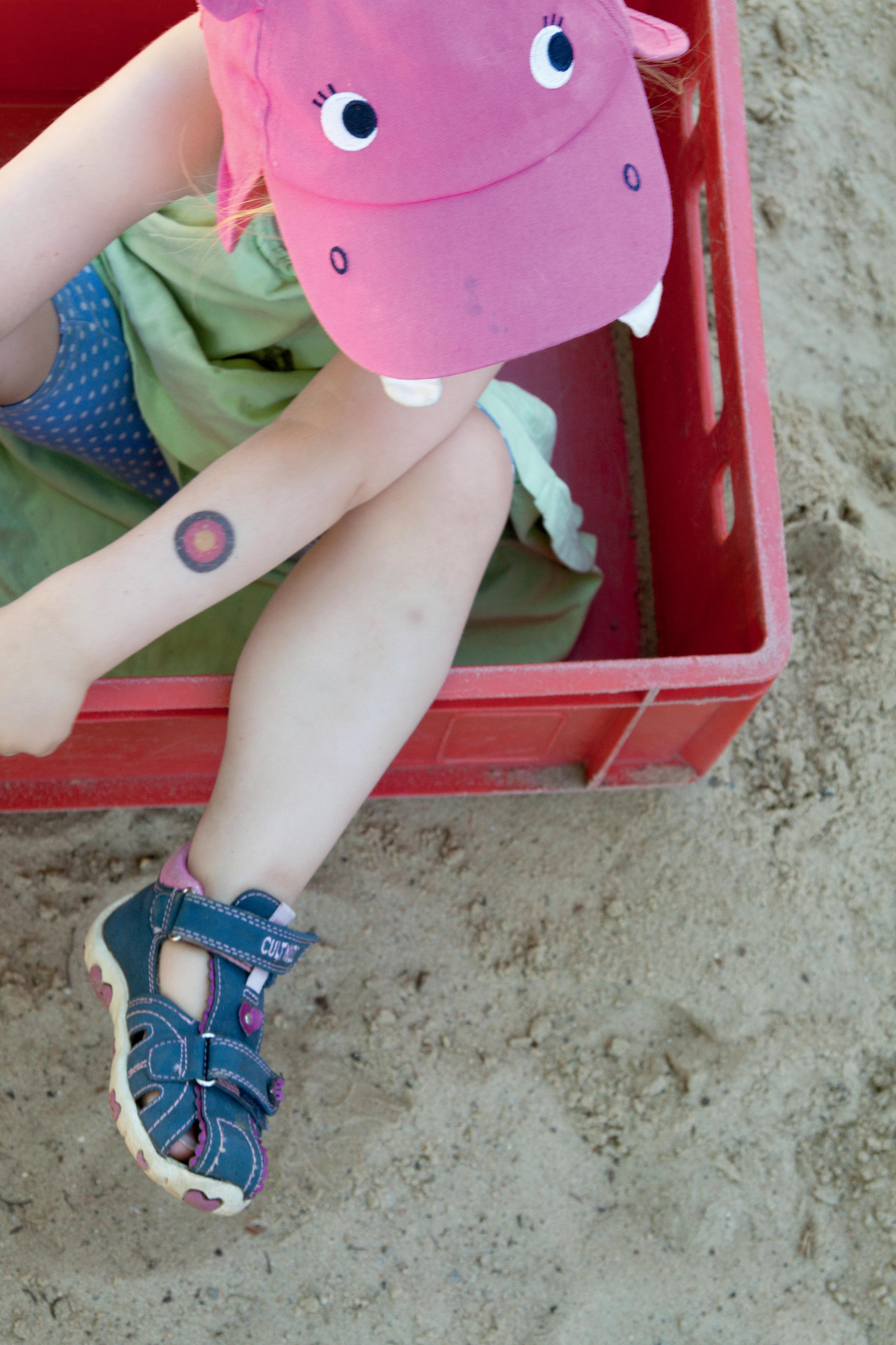 Ein Kind mit einer pinken Mütze sitzt in einer roten Kiste auf einer Sandfläche.
