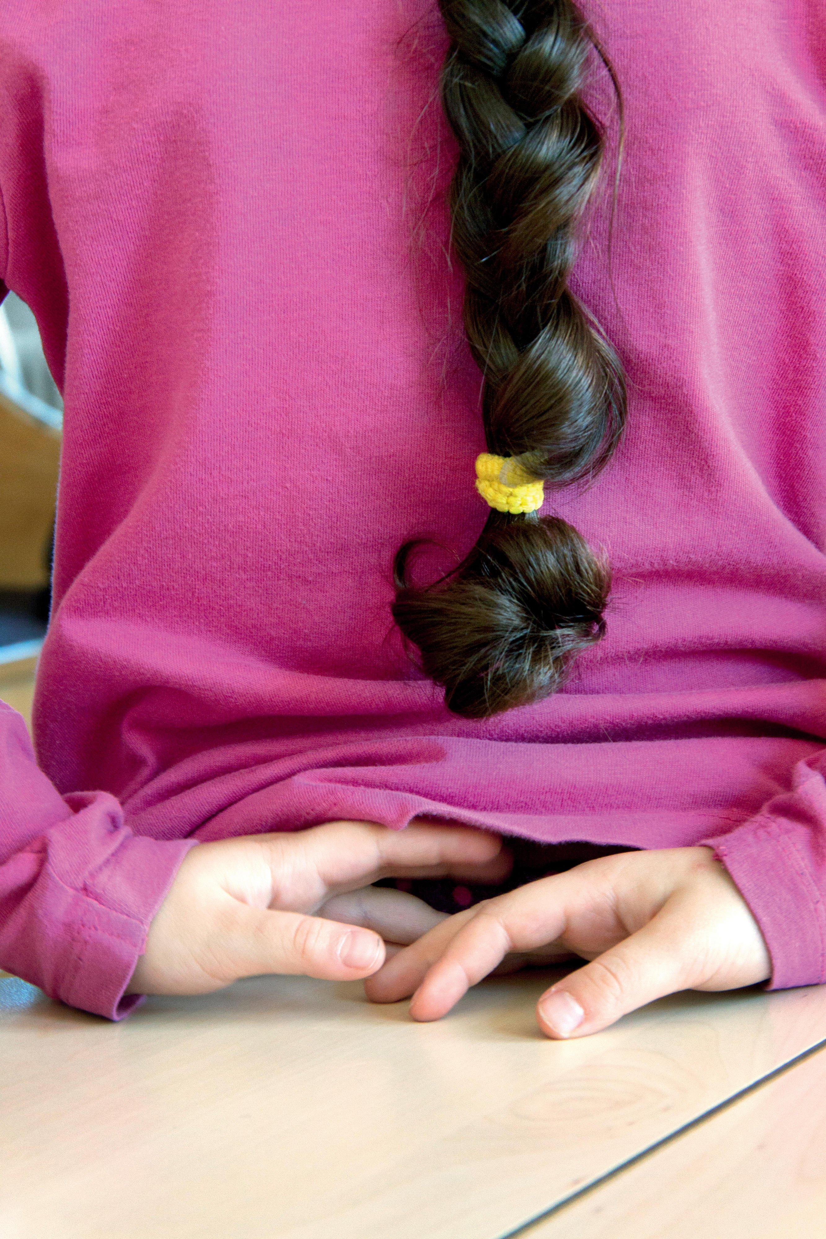 Rückenansicht eines jungen Mädchens in lilanem Pullover mit schwarzem Zopf und hinter dem Rücken verschränkten Händen