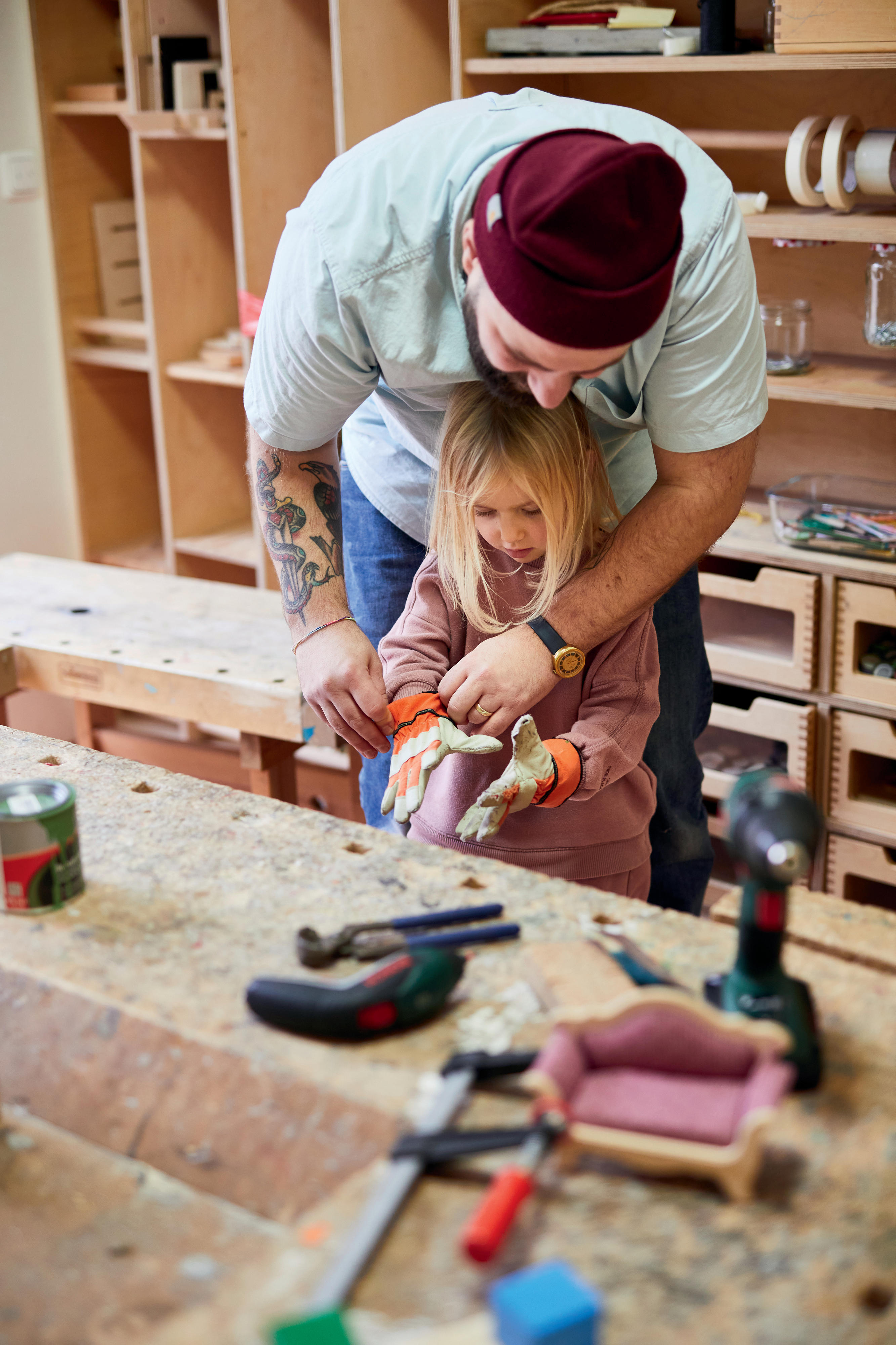 Ein bärtiger Mann mit Wollmütze und Tattoo am Arm hilft einem kleinen Mädchen Arbeitshandschuhe anzuziehen.