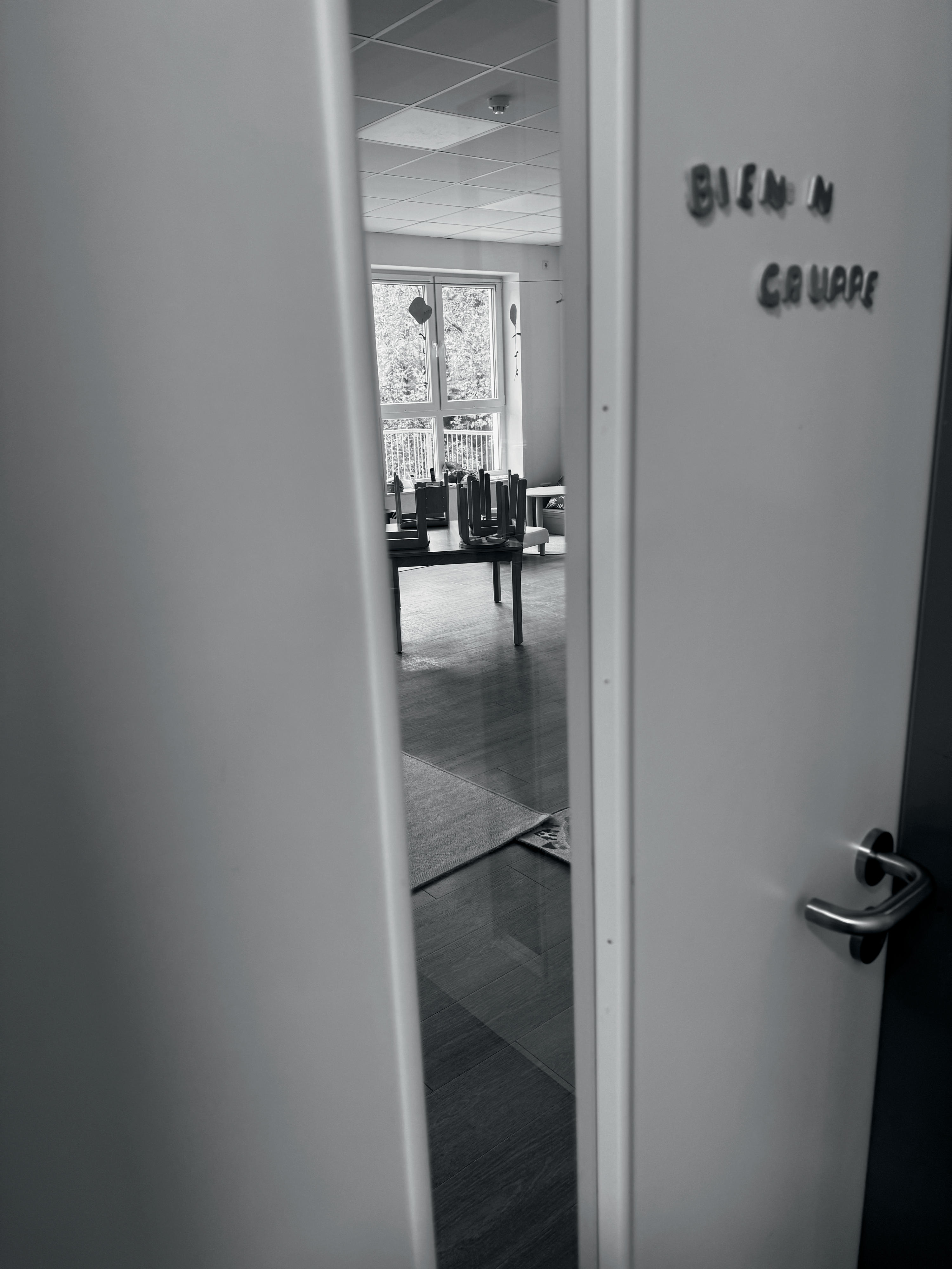 Schwarz-weiß-Bild einer einen Spalt geöffneten Tür zu einem Kita-Raum mit der beschädigten Aufschrift „BIEN N GRUPPE“.
