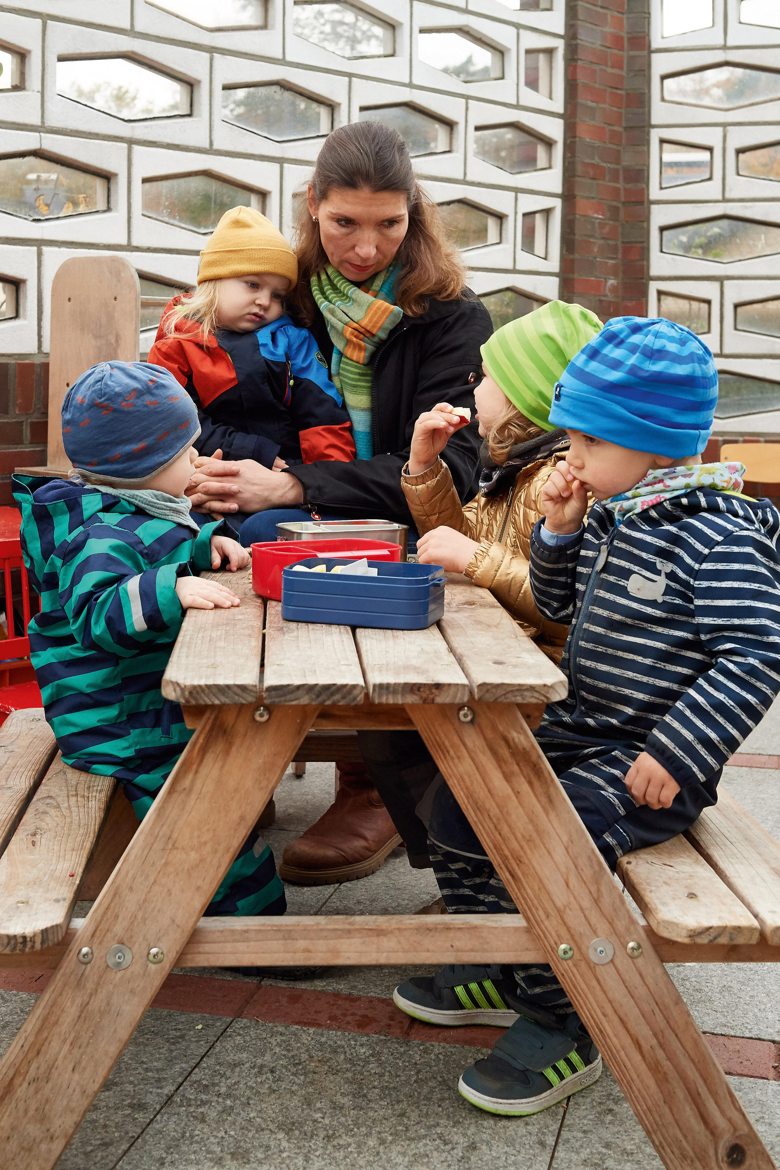 Eine Frau sitzt mit einem Mädchen auf dem Arm draußen am Tisch, daran sitzen noch drei weitere Kinder und essen. Alle tragen Winterkleidung.