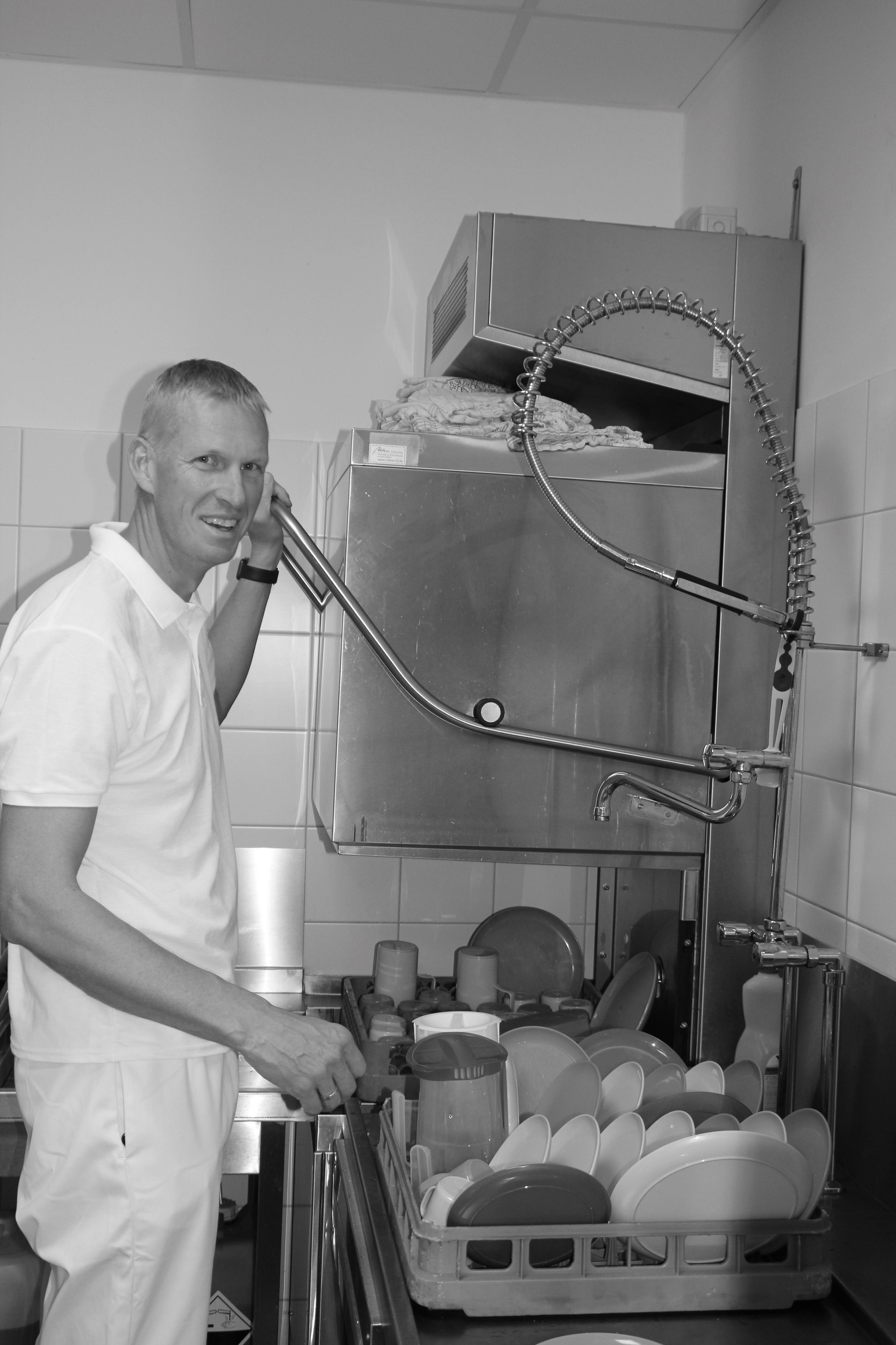 Schwarz-Weiß-Aufnahme: Ein Mann steht vor einer großen Spülmaschine und einem Tablett mit Geschirr.