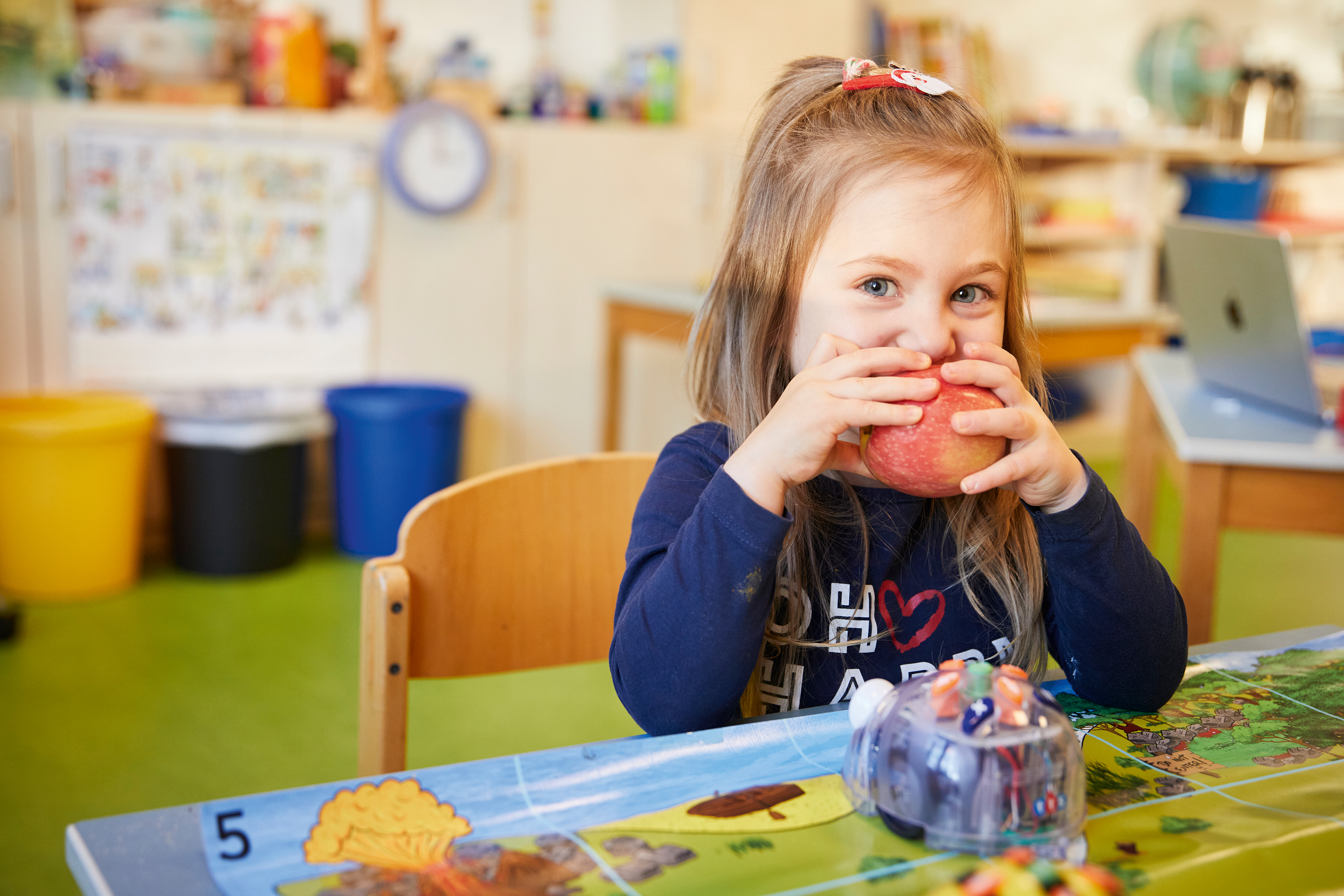 Ein Mädchen sitzt in einer Kita am Tisch und isst einen Apfel, vor ihr steht ein Spielzeugroboter