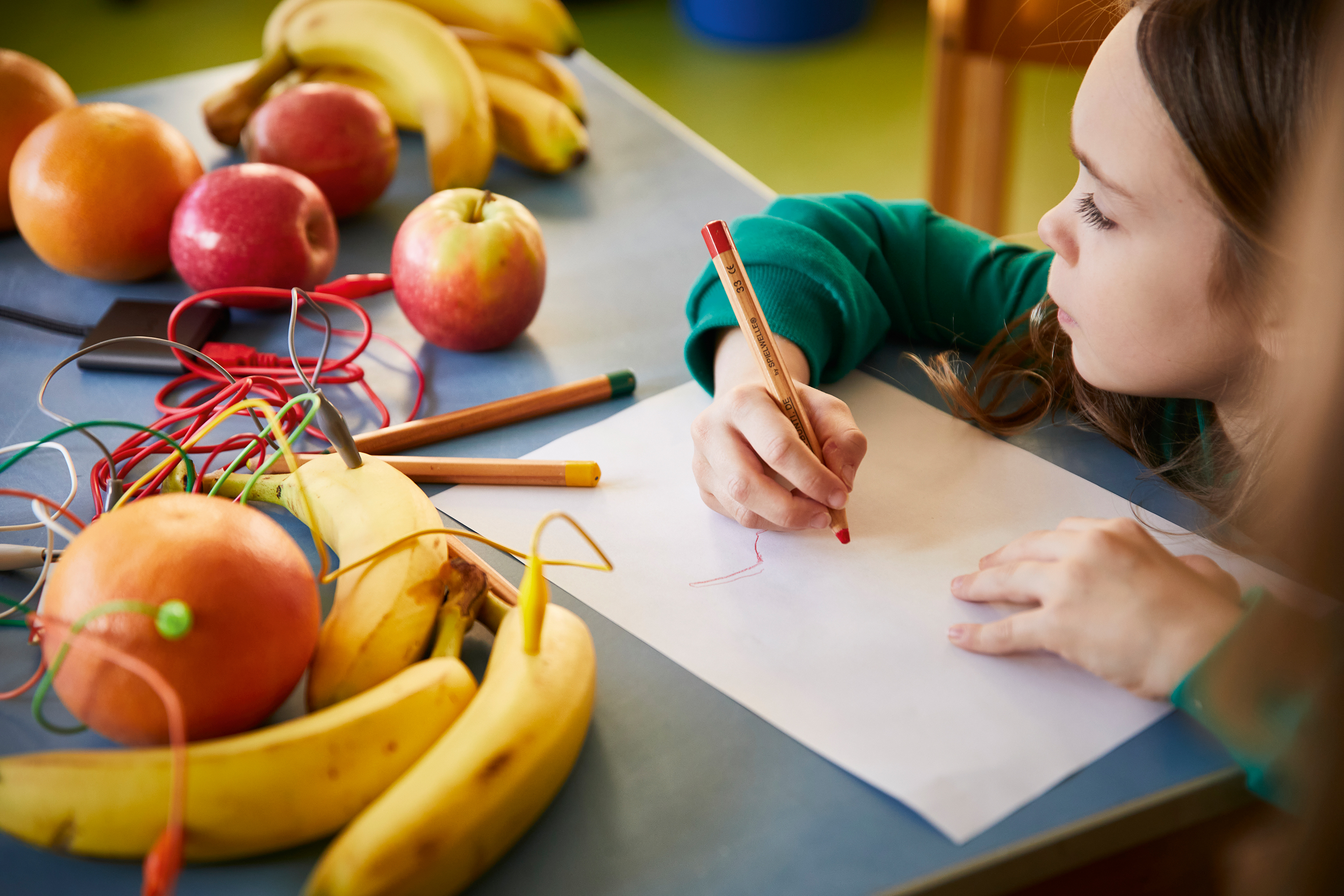 Ein Mädchen sitzt an einem Tisch vor mit Kabeln verbundenen Äpfeln und Bananen und malt.