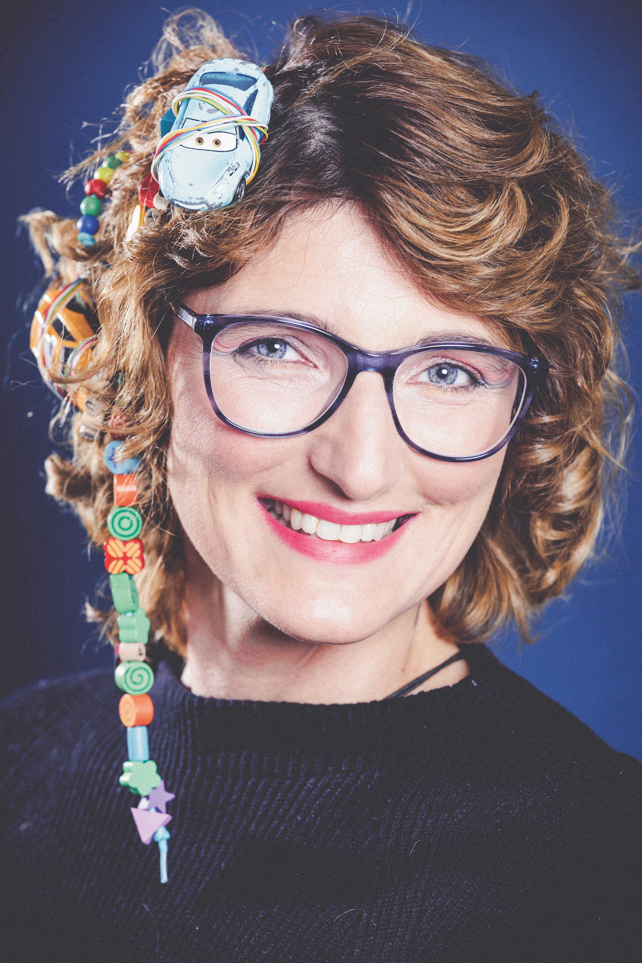 Porträt einer brünetten, lächelnden Frau mit einem Spielzeugauto und bunten Perlenketten im Haar
