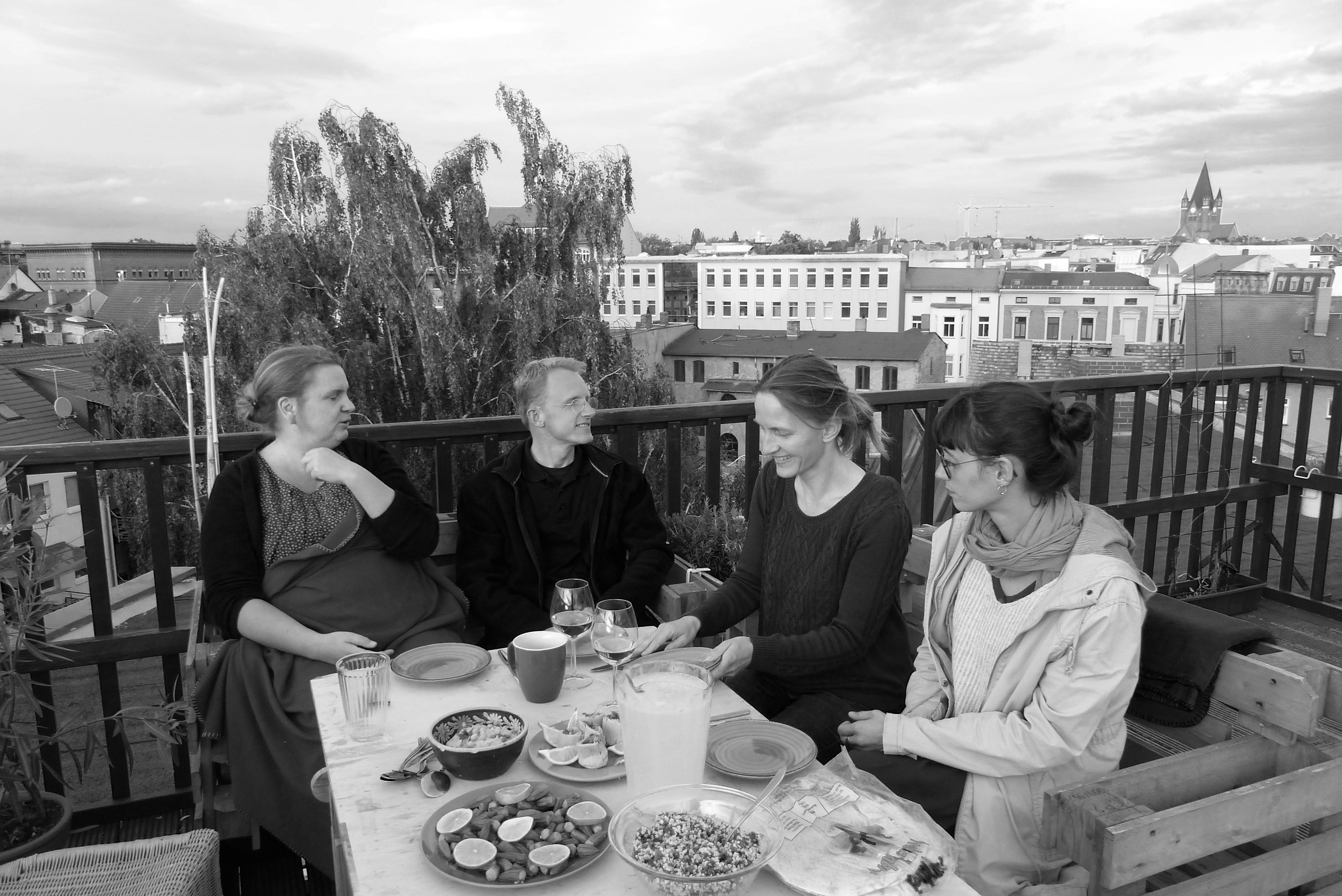 Schwarz-weiß-Aufnahme: Ein Mann und drei Frauen sitzen auf einer Terasse an einem gedeckten Tisch, im Hintergrund ist ein Stadtpanorama zu sehen.