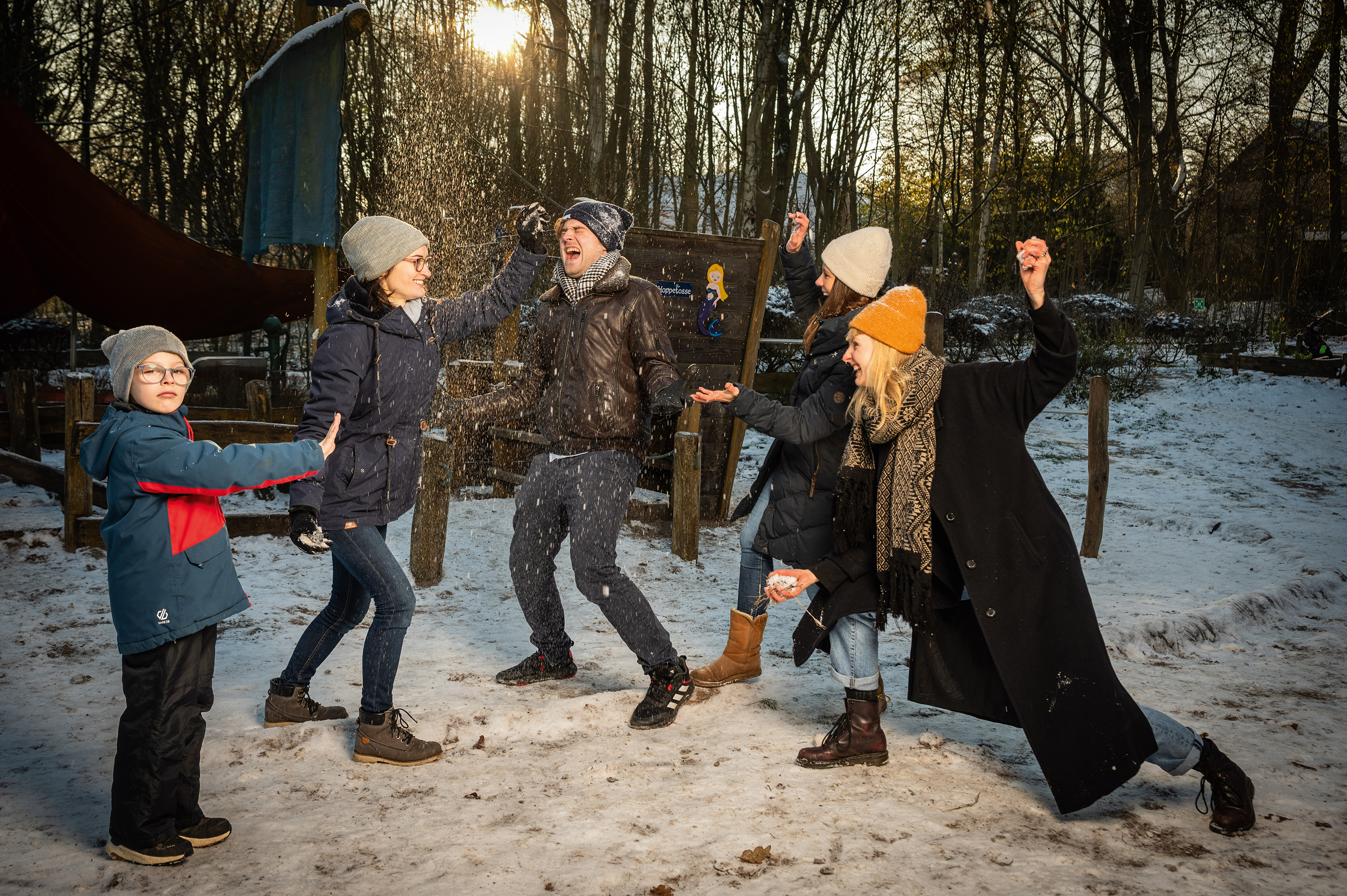 Ein Mann und drei Frauen bewerfen sich gegenseitig mit Schnee, ein Junge steht davor und streckt seine hochgehaltene Hand in ihre Richtung.