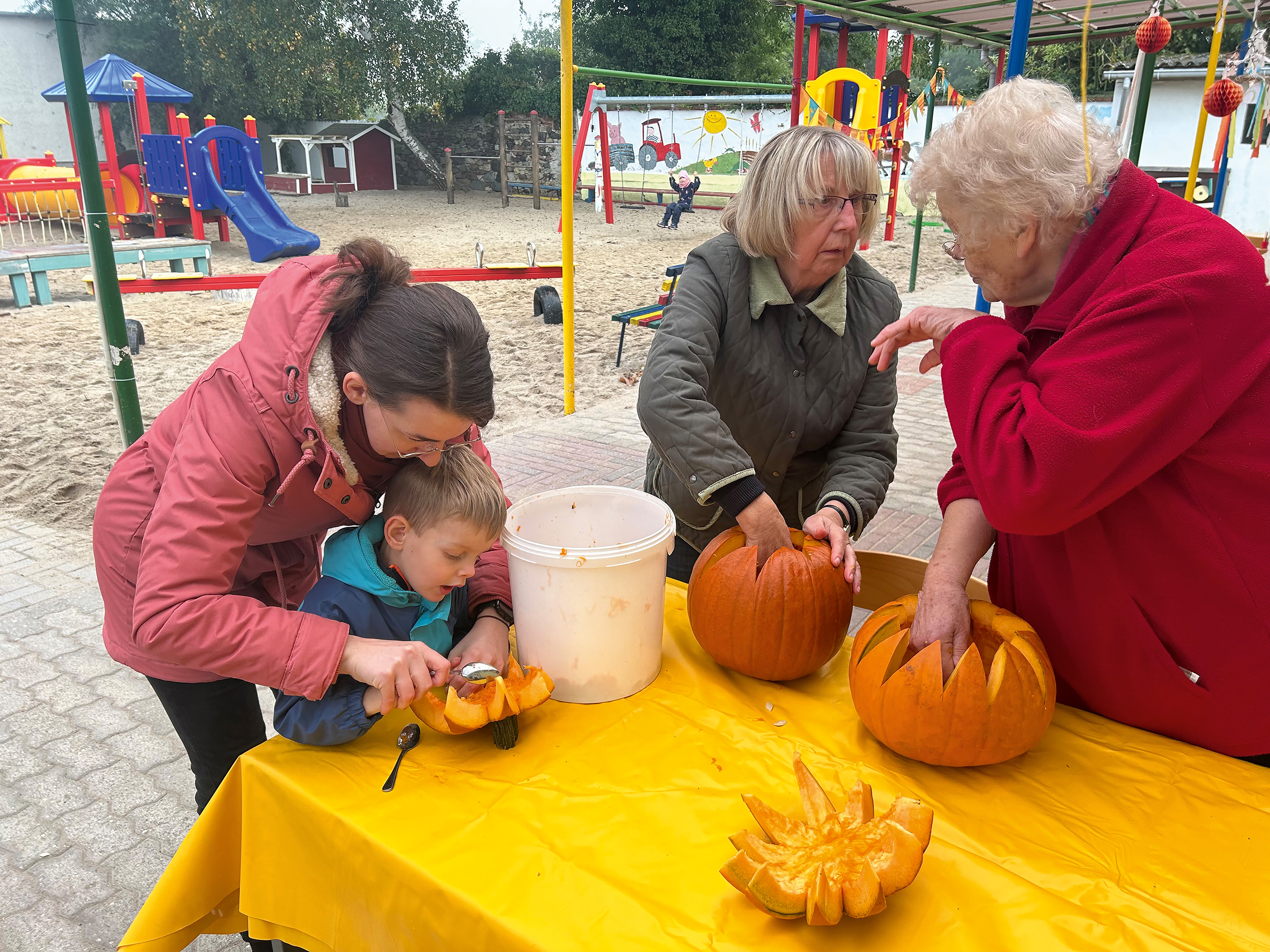 Eine Frau hilft einem Kind dabei an einem Kürbis zuschnitzen, während zwei ältere Frauen sich am selben Tisch unterhalten und auch jeweils einen Kürbis bearbeiten.