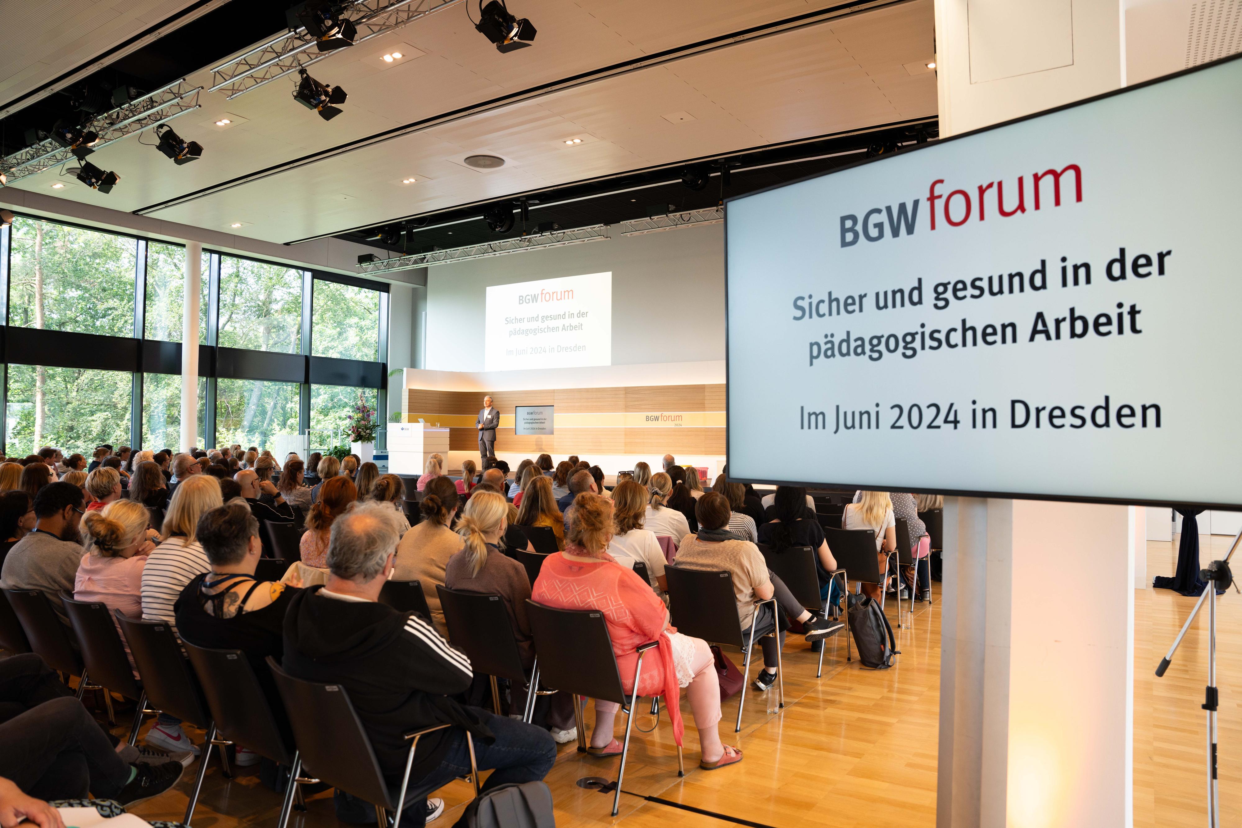 Saalansicht mit sitzendem Publikum. Auf Monitoren steht "BGW forum Sicher und gesund in der pädagogischen Arbeit Im Juni 2024 in Dresden".