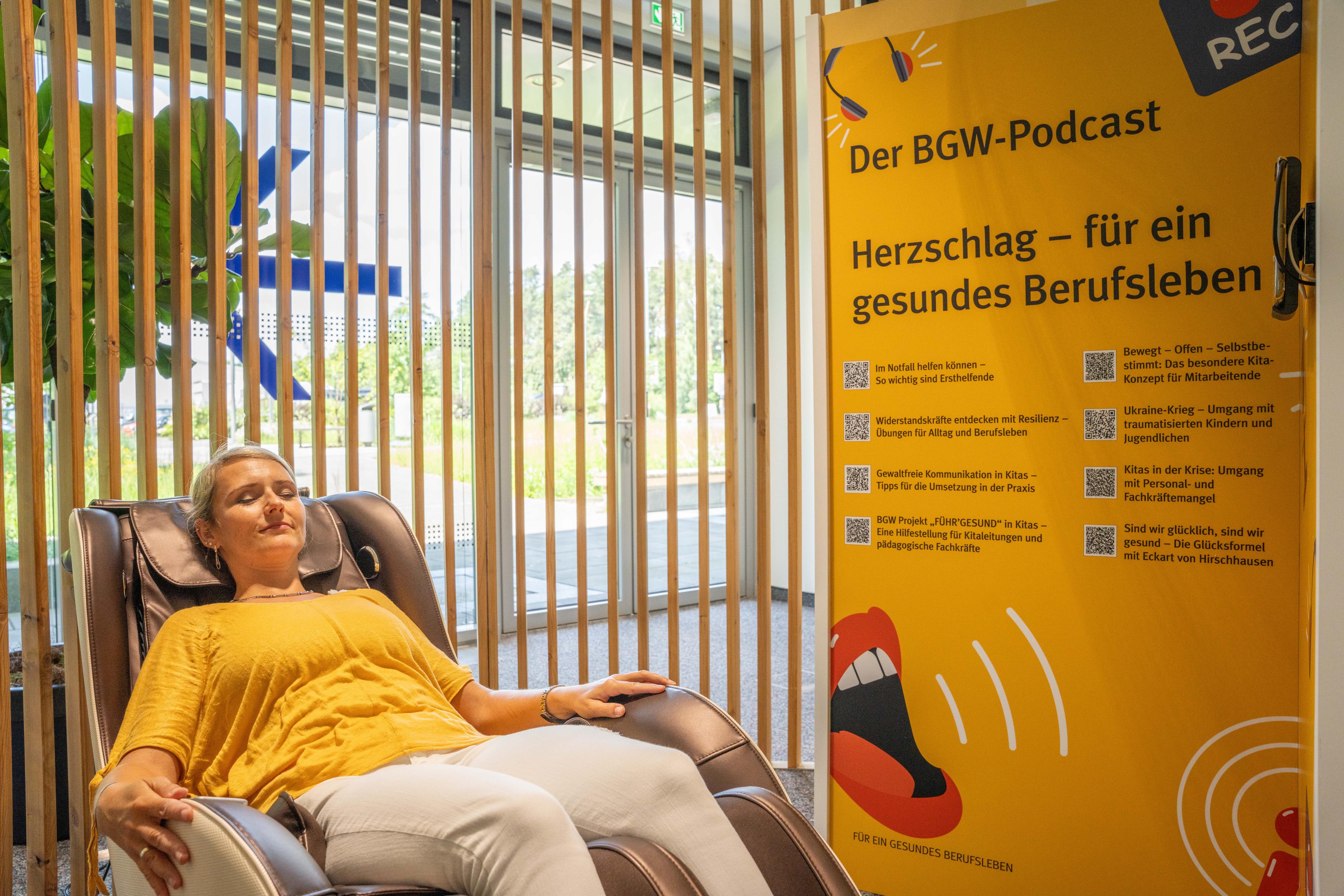 Eine Frau liegt mit geschlossenen Augen in einem Liegesessel, daneben Plakatwand unter anderem mit Text "Der BGW-Podcast. Herzschlag - für ein gesundes Berufsleben"