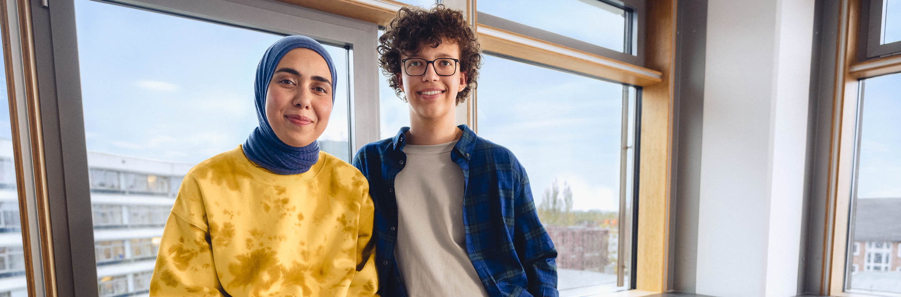 1000 UNDEIN GRUND – Deine Idee von Karriere: mehr bewirken. Eine junge Frau mit Kopftuch und ein junger Mann stehen lächelnd vor einem Fenster in einem Büro.