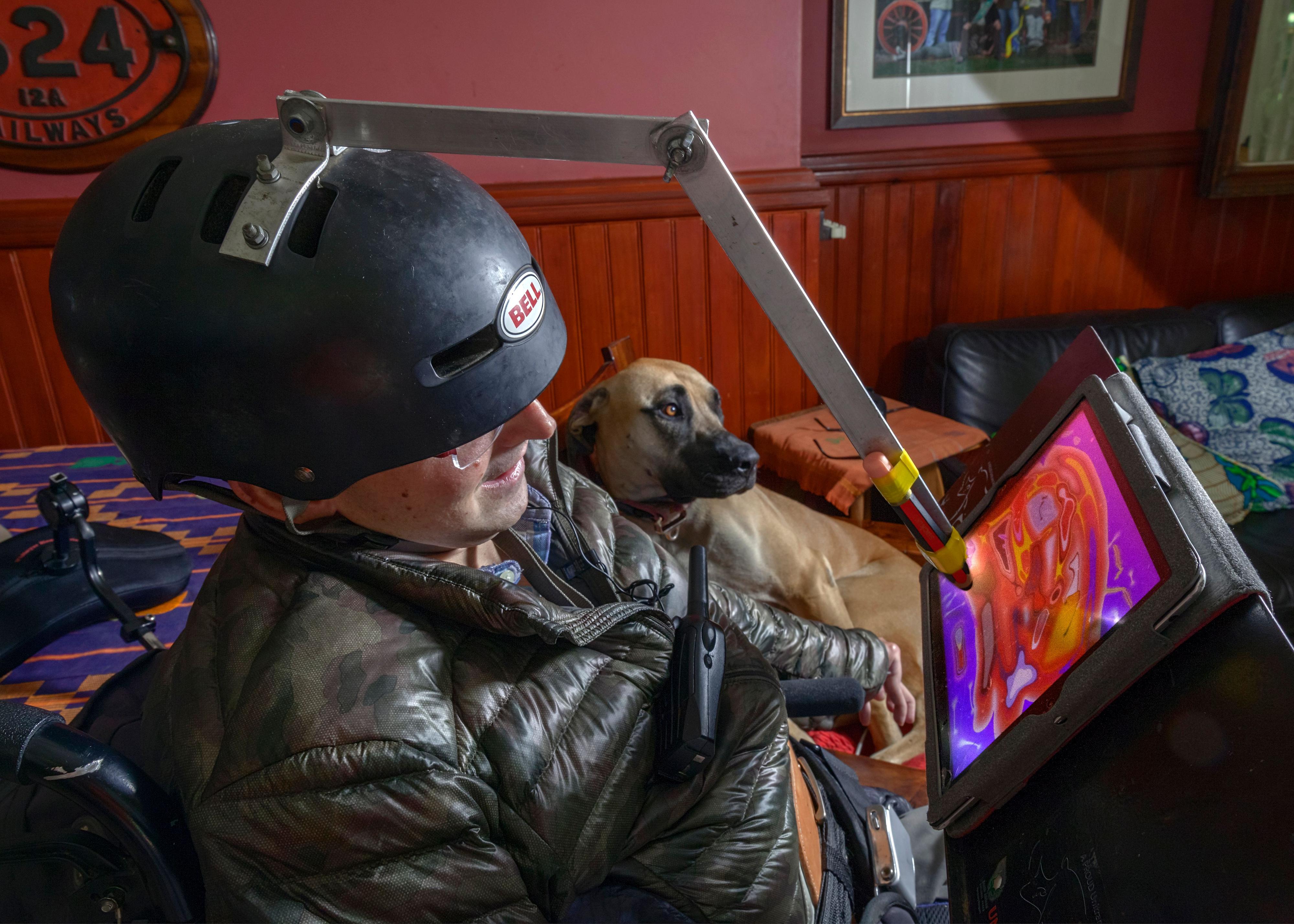 Mensch - Arbeit - Handicap 2021 Ein Mann mit Helm auf dem Kopf. Der Helm hat eine Vorrichtung mit einem Stift, der Farben auf einen Bildschirm malt. Im Hintergrund liegt ein großer brauner Hund.
