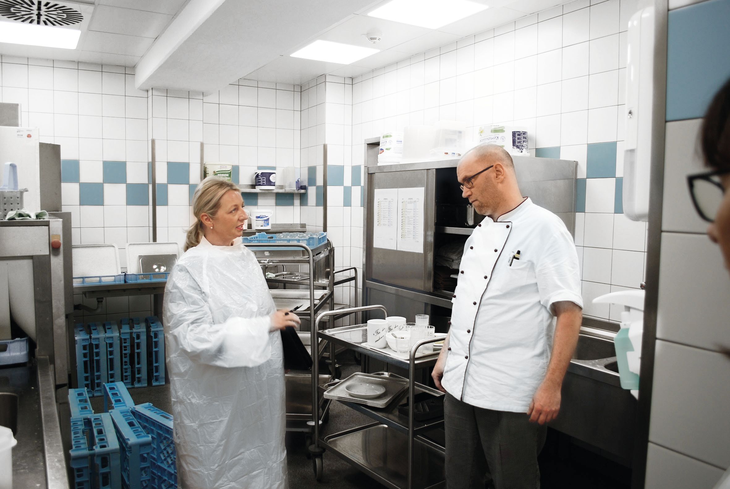 Ruth Giersch spricht mit einem Küchenmitarbeiter neben einem Servierwagen mit Geschirr - sie trägt einen Schutzkittel, er eine Kochjacke.