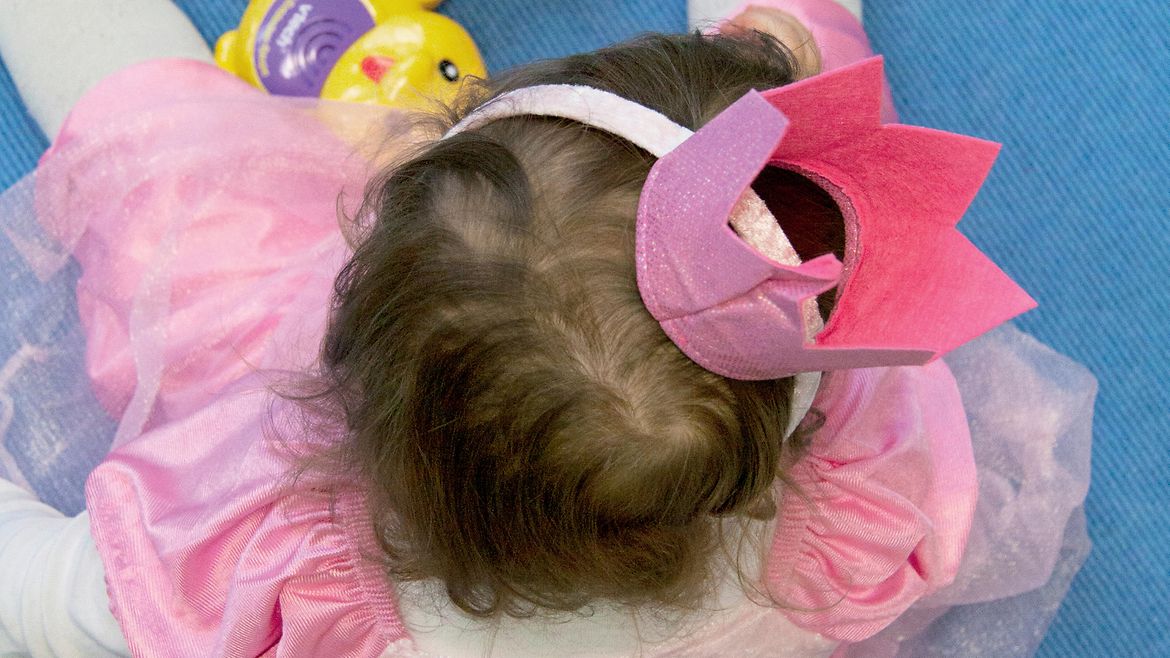 Anischt von oben: Ein Mädchen in pinkem Prinzessinenkostüm mit Krone sitzt auf einem blauen Teppich vor Spielzeug, das Gesicht ist abgewandt.