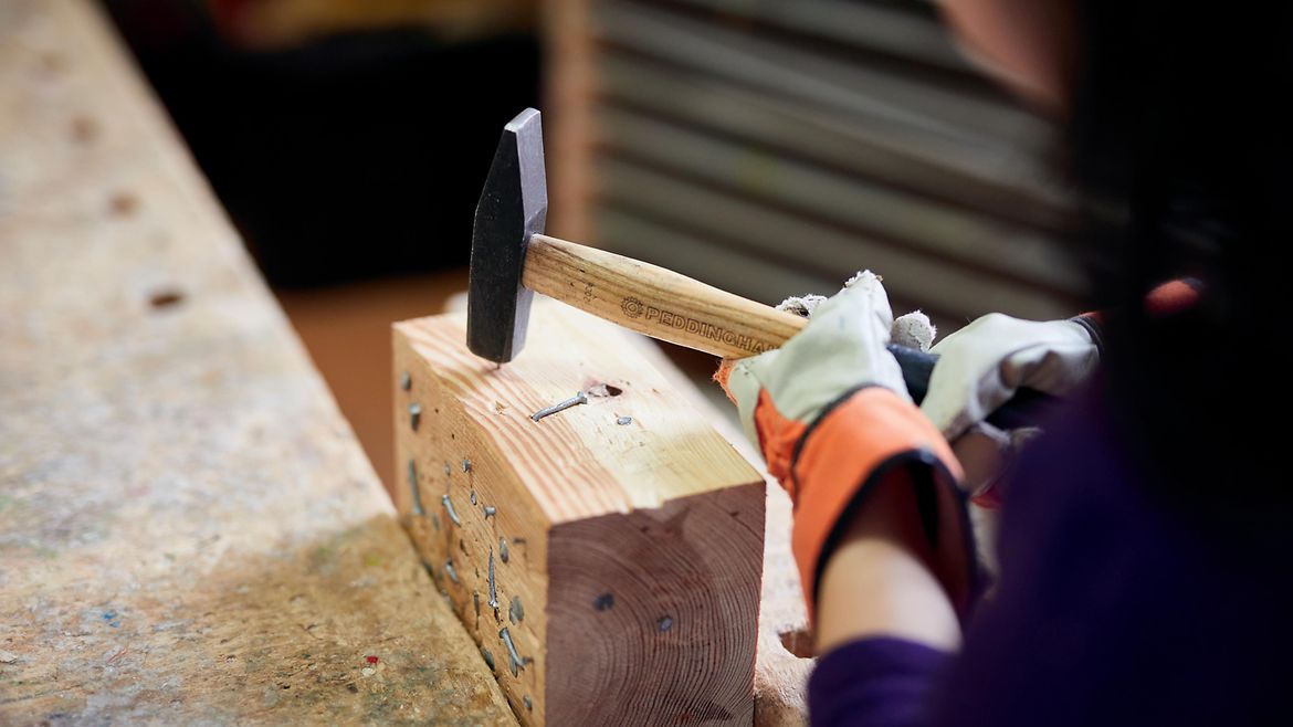 Kinderhände mit Arbeitshandschuhen schlagen mit einem Hammer einen Nagel in ein Stück Holz