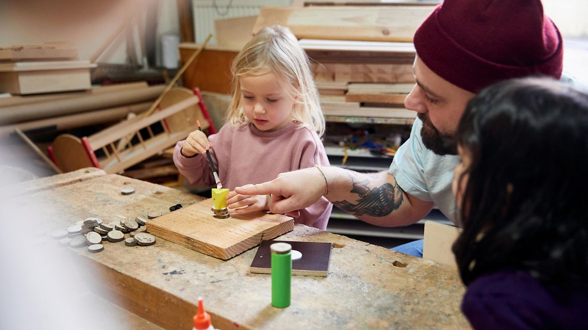 Ein bärtiger Mann mit Wollmütze hilft einem kleinen Mädchen beim Holzwerken