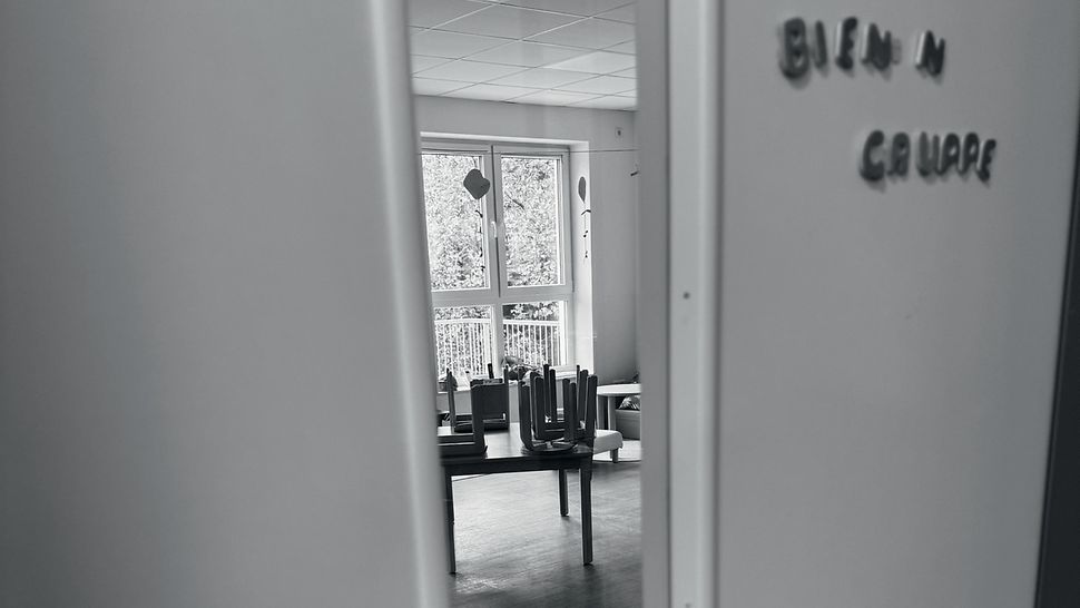 Schwarz-weiß-Bild einer einen Spalt geöffneten Tür zu einem Kita-Raum mit der beschädigten Aufschrift „BIEN N GRUPPE“.