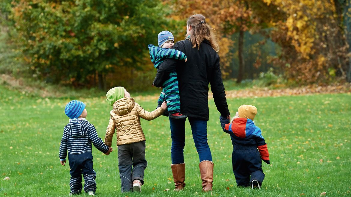 Eine Frau geht mit einem Mädchen auf dem Arm und drei weiteren Kindern, die sich an den Händen halten, über eine herbstliche Wiese. Alle tragen Winterkleidung.