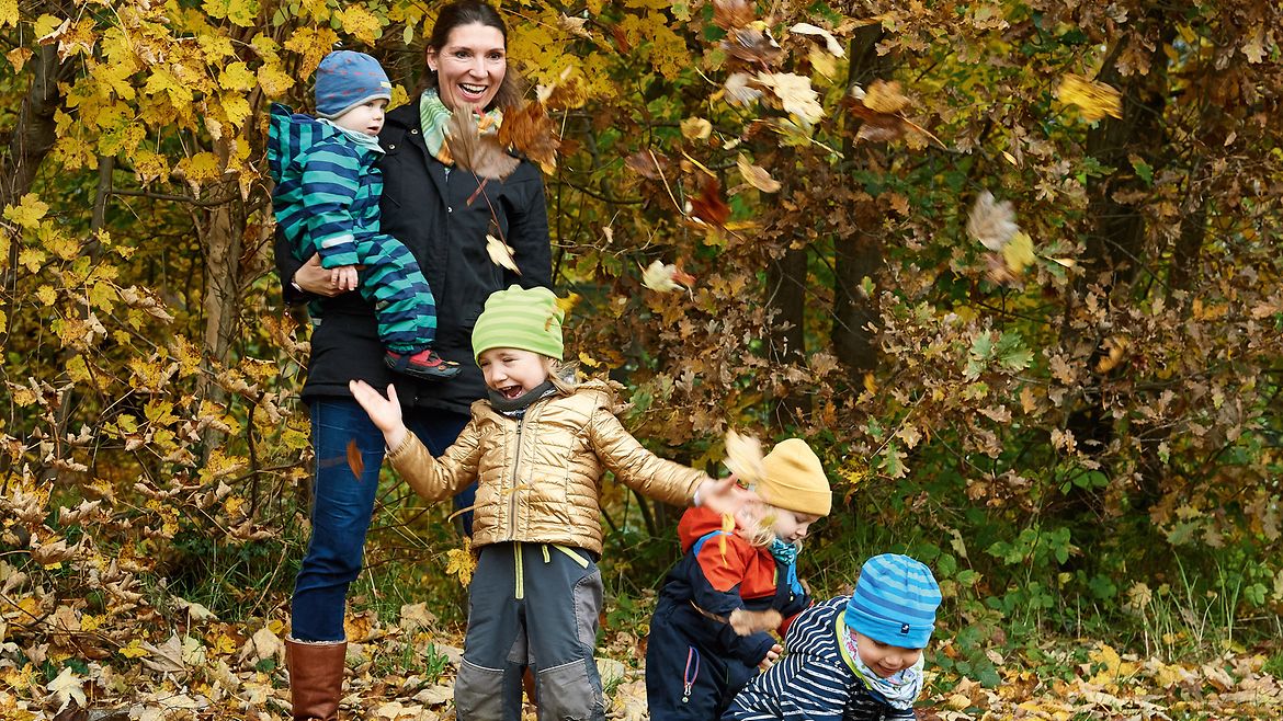 Eine Frau spielt mit vier Kindern im Laub. Alle tragen Winterkleidung.