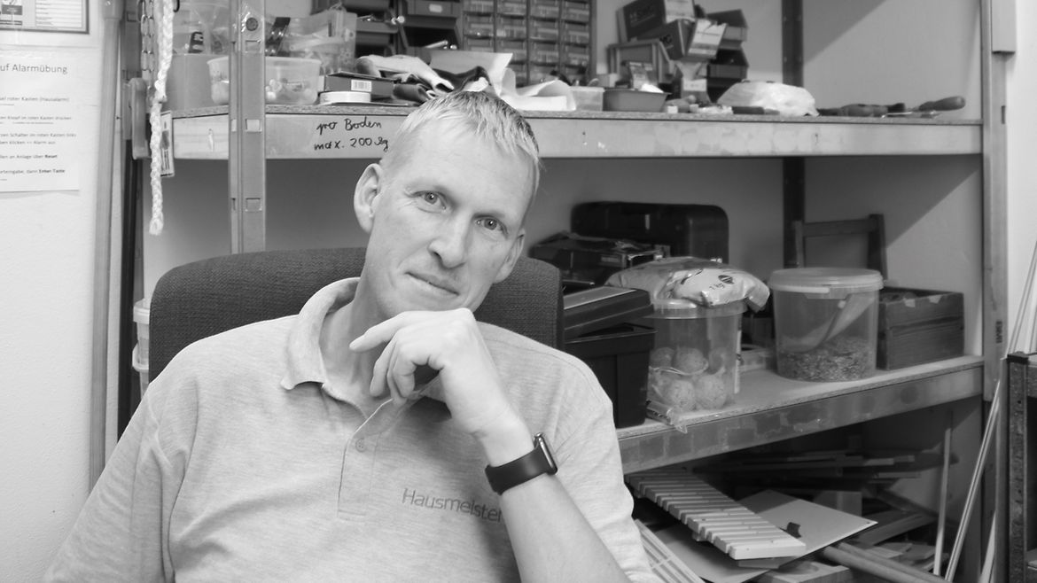 Schwarz-Weiß-Aufnahme: Ein Mann sitzt vor einem Werkzeugregal und schaut freundlich in die Kamera.