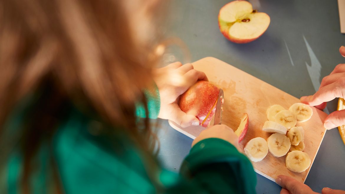 Ansicht von oben: Ein Mädchen schneidet einen Apfel auf einem Brett, daneben liegen Bananenscheiben