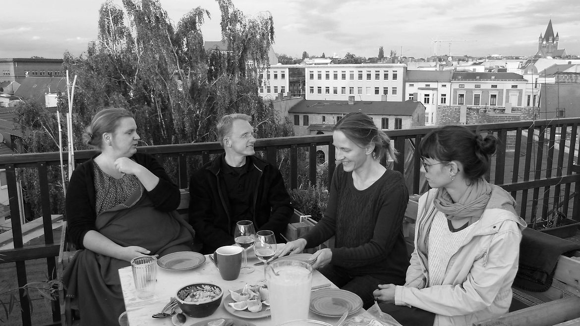 Schwarz-weiß-Aufnahme: Ein Mann und drei Frauen sitzen auf einer Terasse an einem gedeckten Tisch, im Hintergrund ist ein Stadtpanorama zu sehen.