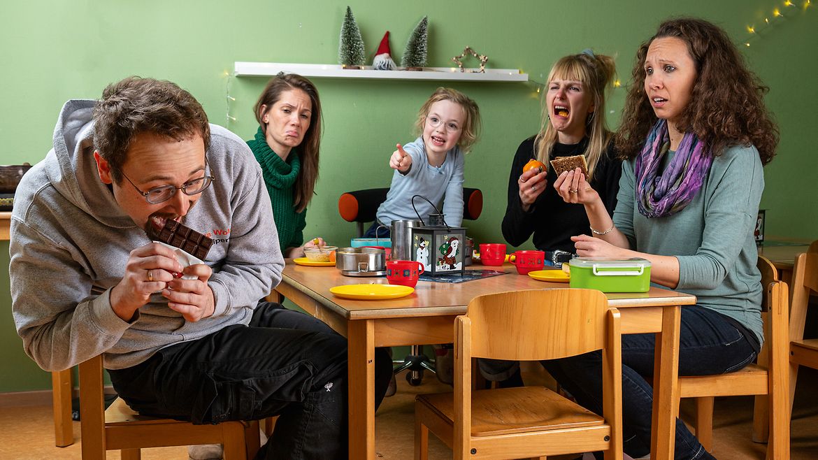 Ein Mann lehnt sich zur Seite und beißt in eine Tafel Schokolade, drei Frauen und ein Kind, die mit ihm am Tisch sitzen, schauen entsetzt, das Kind zeigt mit dem Finger auf ihn.