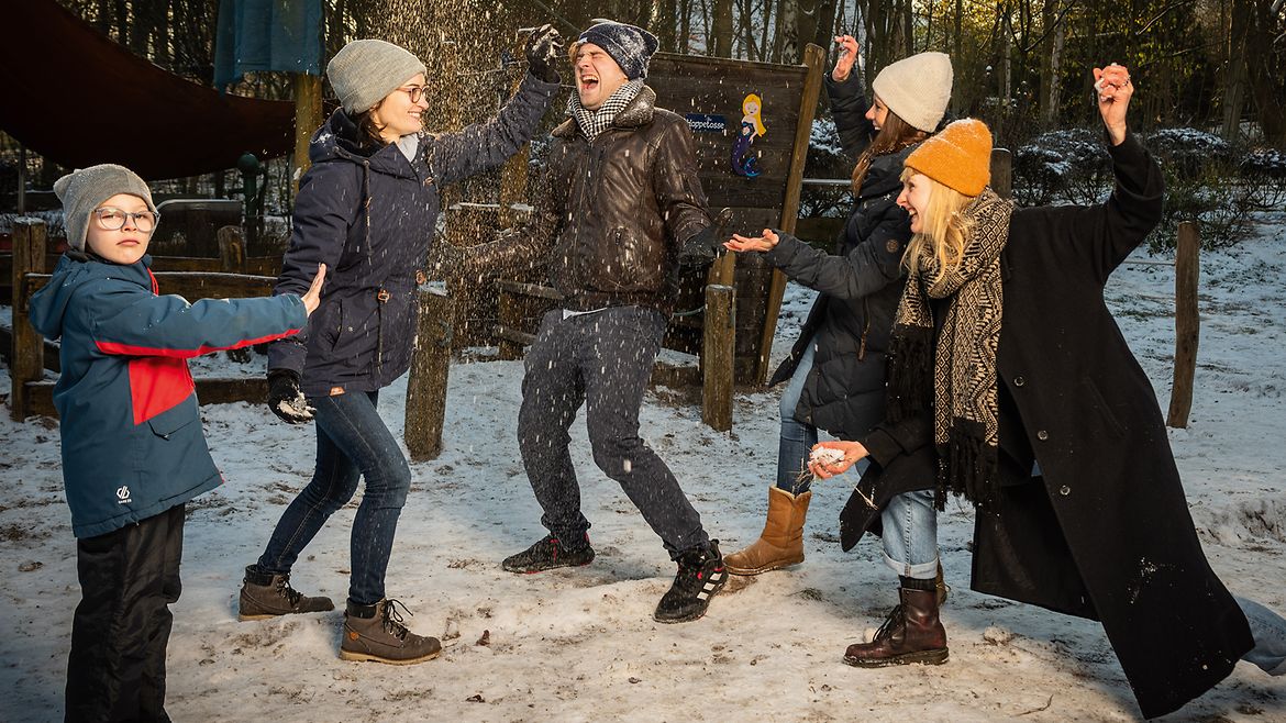 Ein Mann und drei Frauen bewerfen sich gegenseitig mit Schnee, ein Junge steht davor und streckt seine hochgehaltene Hand in ihre Richtung.
