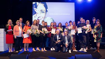 Alle Gewinner des Fotowettbewerbs mit Urkunde und Blumenstrauß in Händen zusammen mit Mitgliedern der Jury auf einer Bühne.
