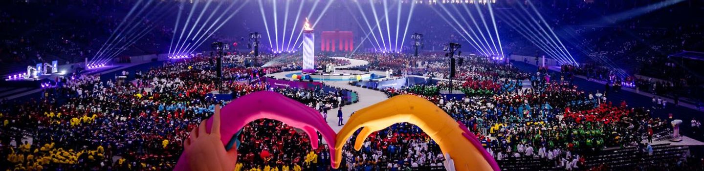 Eröffnungsfeier der Weltspiele im Berliner Olympiastadion: Scheinwerfer und lila Beleuchtung, in der Mitte ein Meer aus Menschen, im Vordergrund zwei Luftschlangenhände, die sich berühren.