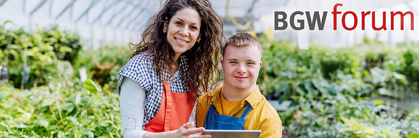 Eine Frau steht neben einem Jungen mit Down-Syndrom, der ein Tablet in der Hand hält, in einem Gewächshaus vor grünen Pflanzen. Beide tragen Arbeitskleidung und lächeln in die Kamera. Oben links steht der Schriftzug "BGW forum".