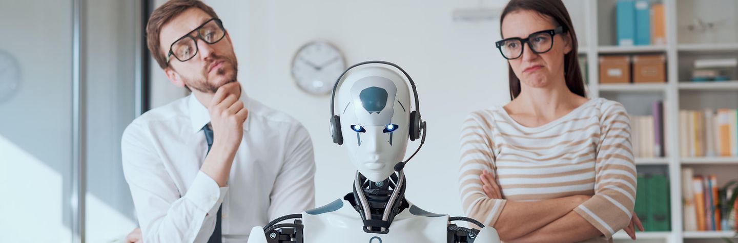 Ein Roboter sitzt mit Headset vor einem Laptop am Schreibtisch in einem Büro. Dahinter stehen ein Mann und eine Frau, die ihn skeptisch bis