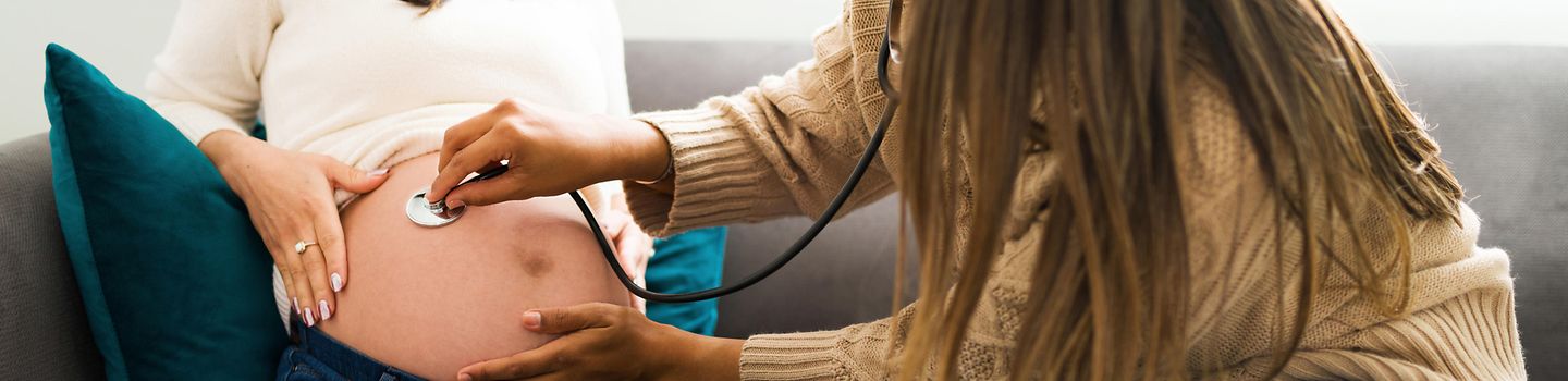 Hebamme untersucht schwangere Frau Hebamme benutzt Stethoskp auf dem Bauch schwangeren Frau. Beide sitzen auf einem Sofa in einem Wohnzimmer.