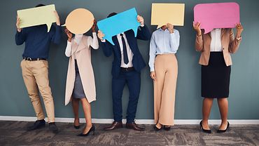 Anonyme Personalbefragung Drei Frauen und zwei Männer stehen nebeneinander vor einer Wand und halten Sprechblasen aus bunter Pappe vor ihre Gesichter.